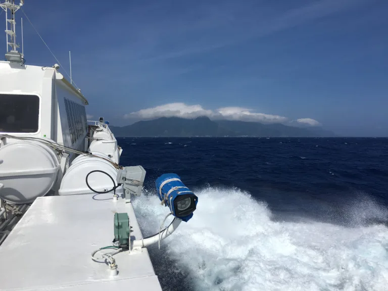White passenger boat cruising at sea with a wake splashing at the bow and distant cloud-covered mountains on the horizon.